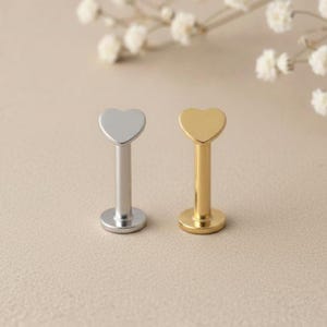 Puede incluir: Dos piercings labiales. Uno es plateado y el otro dorado. Cada uno tiene una parte superior en forma de corazón y un poste recto. Los piercings se muestran sobre un fondo neutro con iluminación suave, lo que sugiere un producto de joyería.