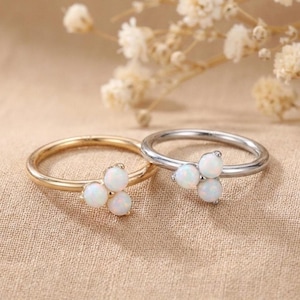 May include: Two rings, one gold and one silver, each featuring three white opal stones clustered together. The rings are set against a neutral background with delicate white flowers, showcasing the jewelry's elegant design.