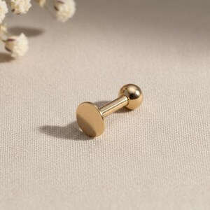 May include: A gold-colored, flat-backed earring with a small, round ball on one end. The earring is a simple, minimalist design, perfect for everyday wear. The earring is made of a shiny, metallic material.