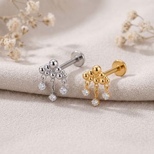 May include: Two labret piercings featuring a cloud-shaped top with three dangling chains, each ending in a small clear round gemstone. One piercing is silver-toned metal, and the other is gold-toned metal, both with a flat back post design.