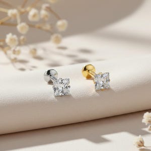 May include: Two stud earrings, one silver and one gold, each featuring a square, clear gemstone. The earrings are displayed on a light-colored surface, with soft, blurred floral accents in the background. The earrings are designed for pierced ears.