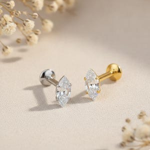May include: Two stud earrings with marquise-cut clear gemstones. One earring has a silver post and setting, while the other is gold. The earrings are displayed on a neutral-toned surface, with soft lighting and floral accents.