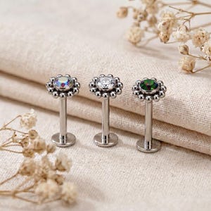 May include: Three silver-toned labret studs featuring a flower-shaped head with a central gemstone. The left stud has an iridescent stone, the middle has a clear stone, and the right has a dark green stone, all surrounded by small metal beads.