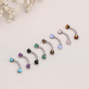 May include: A collection of curved eyebrow rings with various gemstone beads. The jewelry features a silver-colored, curved barbell design with gemstones in shades of turquoise, amethyst, black, green, brown, light blue, pink, and tiger's eye.