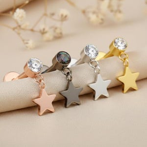 May include: Four stud earrings with star charms. Each earring features a different metal finish: rose gold, gunmetal, silver, and gold. The studs have a clear, round gemstone. The star charms dangle below the gemstone.