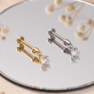 May include: Two pieces of jewelry, one gold and one silver, each with a long post and a dangling pair of clear, round gemstones. The jewelry is displayed on a reflective surface.