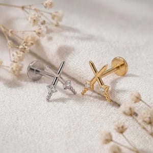 May include: Two flat back stud earrings shaped like crossed swords. One earring is silver-toned metal with a clear gemstone at the hilt, and the other is gold-toned metal with a clear gemstone at the hilt. Both feature a post and a circular backing.