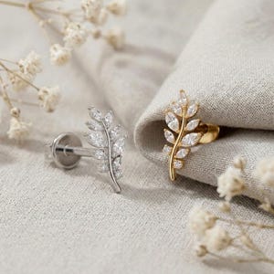May include: Two stud earrings shaped like leaves, one in silver tone and one in gold tone. Each earring features a central stem with multiple marquise-cut clear stones arranged in a branch pattern, with a flat circular backing post visible.