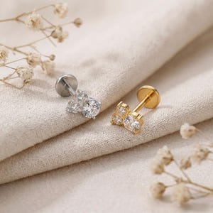 May include: Two flat back stud earrings, one in silver tone and one in gold tone, each featuring a cluster of three round clear cubic zirconia stones arranged in a triangular pattern with a circular base post for secure placement in the ear.