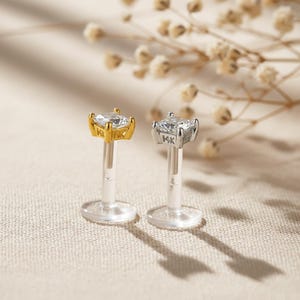 May include: Two labret studs with clear cubic zirconia stones. One stud has a gold setting marked "14K", the other has a silver setting also marked "14K". Both have clear posts and flat, round bases.