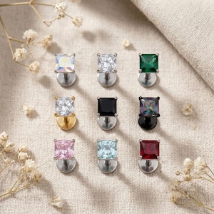 May include: A collection of nine square-cut stud earrings arranged in a grid. The earrings feature various colored stones including iridescent, clear, emerald green, black, rainbow, pink, light blue, and deep red, set on metallic posts.