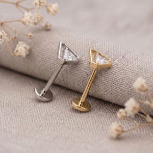 May include: Two labret studs featuring a triangular head with a clear, faceted stone set inside a metal frame. One stud is silver-toned and the other is gold-toned, both with a flat circular back disc and a straight post for secure placement.