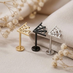May include: Three labret studs featuring a flat circular base, a straight post, and a top shaped like a spider web. The set includes one gold-toned, one black, and one silver-toned stud, each with a cutout web design on the top portion.