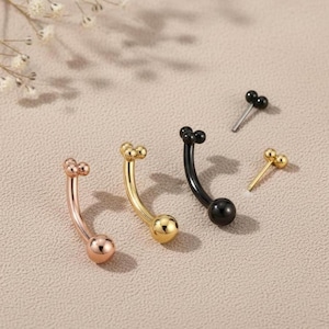 May include: An assortment of curved barbell piercings in various colors. The piercings include rose gold, gold, and black, each with a ball at the bottom and two or three smaller balls at the top. A small silver piercing with black balls is also present.