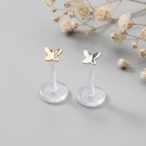 May include: Two butterfly-shaped lip or cartilage studs. One is gold-colored, the other silver. Both have clear, plastic posts and circular bases. The background is a soft gray with dried flowers.