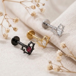 May include: Three flat-back labret studs featuring a heart-shaped frame with thorn-like details. One stud is black with a red teardrop-shaped stone, one is gold with a clear teardrop stone, and one is silver with a clear teardrop stone.