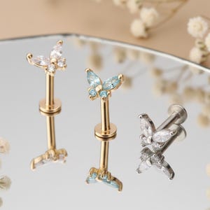 May include: Three butterfly-shaped earrings. Two are gold-toned with clear and light blue stones, and one is silver-toned with clear stones. The earrings are displayed on a reflective surface.
