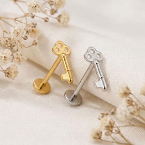 May include: Two flat back stud earrings shaped like skeleton keys. One earring is gold-toned and the other is silver-toned. Each stud features a circular head with cutouts, a straight post, and a flat circular base for secure placement.