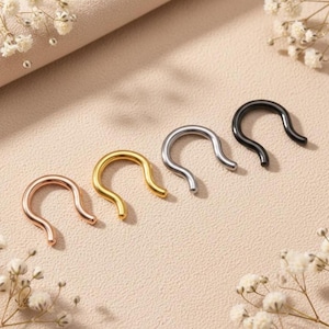 May include: Four nose rings in rose gold, gold, silver, and black. The U-shaped rings are displayed on a neutral surface, with small white flowers in the background. These are a type of body jewelry.