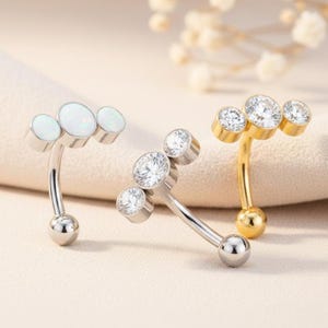 May include: Three curved belly button rings. One is silver with three white opal stones. Another is silver with three clear, sparkling stones. The third is gold with three clear, sparkling stones. Each ring has a small ball at the bottom.