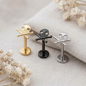May include: Three labret body jewelry pieces featuring a skull and crossbones design. The set includes one gold-tone, one black, and one silver-tone stud, each with a flat circular base and a vertical post topped with a skull and crossbones motif.