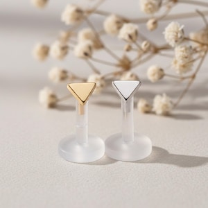 May include: Two lip piercing studs. One stud has a gold-colored triangle top, and the other has a silver-colored triangle top. Both studs have a clear, plastic post and a clear, round base. The background includes white flowers.