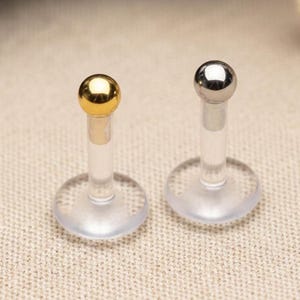 May include: Two lip piercing studs. One has a gold-colored ball, the other a silver-colored ball. Both have a clear, straight post and a clear, round base. The background is a neutral beige.