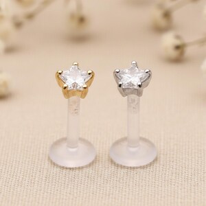 May include: Two star-shaped lip piercing studs. One stud has a gold-colored setting, and the other has a silver-colored setting. Both have a clear, star-shaped gem and a clear post with a flat disc backing.