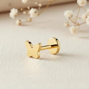 May include: A gold-colored butterfly stud earring. The earring features a butterfly design on the front and a flat disc on the back. The earring is set against a soft, neutral background with delicate white flowers.