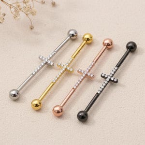 May include: Four industrial barbell earrings in silver, gold, rose gold, and black. Each barbell features a cross design encrusted with clear crystals. The earrings have a ball on each end.