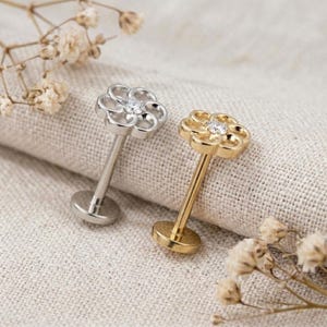 May include: Two labret studs featuring a flower-shaped top with a central clear stone. One stud is silver-toned and the other is gold-toned. Each piece has a flat circular base and a straight post, designed for body piercing jewelry applications.