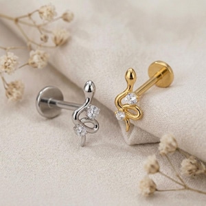 May include: Two flat back labret studs featuring a snake-shaped design. One stud is silver-toned with two clear round stones, and the other is gold-toned with two clear round stones. Both pieces have a straight post and a circular flat back base.