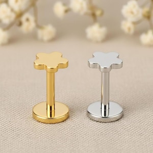 May include: Two flower-shaped lip piercing studs. One is gold-colored, the other is silver-colored. Each stud has a flat, round base and a post that connects to the flower-shaped top. The background is a neutral color.