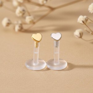 May include: Two lip piercing studs. One has a gold heart-shaped top, the other a white heart. Both have clear posts and flat, round bases. The background is a soft, neutral color with blurred floral accents.