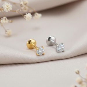 May include: Two small stud earrings, one gold-toned and one silver-toned, each featuring a clear, round gemstone. The earrings are displayed on a soft, neutral-colored fabric, with delicate white flowers in the background.
