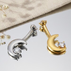 May include: Two belly button rings, one silver-toned and one gold-toned, each featuring a crescent moon design. The moon shape cradles a clear, round gemstone. The jewelry is displayed on a reflective surface.
