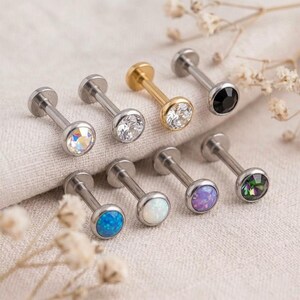 May include: A collection of seven labret studs featuring flat back posts and circular tops. The tops include clear crystals, a black stone, a gold-toned metal setting, and various iridescent, blue, purple, and green opal-style cabochons.