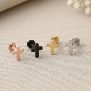 May include: Four cross-shaped stud earrings in rose gold, black, gold, and silver. The earrings are displayed on a neutral surface, showcasing their different metallic finishes and simple design. These earrings are suitable for various styles.
