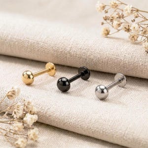 May include: Three labret studs with flat back discs and spherical ends, arranged in a row. The studs are finished in metallic gold, glossy black, and polished silver tones, each featuring a smooth, rounded ball top and a straight post.