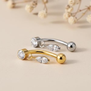May include: Two curved belly button rings. One is gold-toned with a clear, round gem and a dangling marquise-cut gem. The other is silver-toned with a similar dangling gem. Both have a ball on the end.