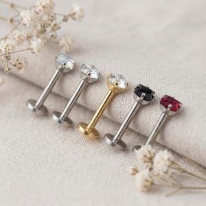 May include: Five labret studs with flat back discs and prong-set stones. Two studs have silver-tone metal with clear stones, one has gold-tone metal with a clear stone, one has silver-tone metal with a black stone, and one has silver-tone metal with a red stone.