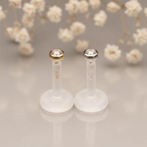 May include: Two lip piercing studs. One has a gold-colored bezel setting, and the other has a silver-colored bezel setting. Both have a clear, round base and a clear post. The studs are set against a soft, blurred background.