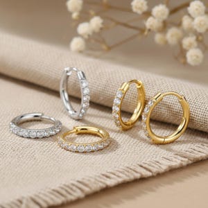 May include: An assortment of hoop earrings in silver and gold tones, each adorned with a row of small, clear gemstones. The earrings are displayed on a neutral-colored fabric, with soft lighting and a blurred floral background.