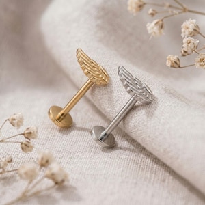 May include: Two labret piercings featuring wing-shaped tops, one in a polished gold tone and the other in a polished silver tone. Each piece consists of a flat circular base, a straight post, and a detailed wing design on the front-facing end.