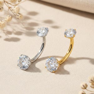 May include: Two belly button rings. One is silver-toned, the other is gold-toned. Each ring features a curved bar with a large, clear, round gemstone at each end. The gemstones sparkle, reflecting light. The background is a soft, neutral color.