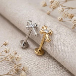 May include: Two flat back labret studs featuring cross-shaped tops. One stud is silver-toned with a clear round gemstone at the center of the cross, and the other is gold-toned with a matching clear round gemstone at the center of the cross.