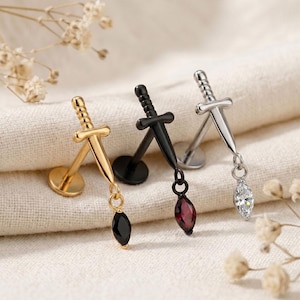 May include: Three labret stud earrings featuring a sword-shaped charm. One is gold-toned with a black marquise-cut stone, one is black with a dark red marquise-cut stone, and one is silver-toned with a clear white marquise-cut stone.