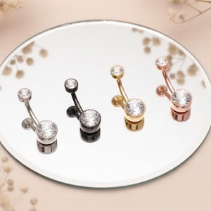 May include: Four belly button rings in silver, black, gold, and rose gold. Each ring features a large, clear, round gemstone at the bottom and a smaller gemstone at the top. The rings are displayed on a reflective surface.