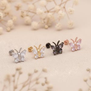 May include: Four butterfly-shaped stud earrings in silver, gold, black, and rose gold. Each earring features a butterfly design with clear or iridescent stones. The earrings are displayed on a light beige surface.