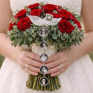 Puede incluir: Un ramo de novia de rosas rojas intensas y follaje verde con un lazo de cinta blanca y una cadena vertical de dijes plateados con cinco fotografías circulares de retratos en blanco y negro que cuelgan del centro.